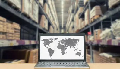 Laptop showing a world map in a warehouse with shelves of boxes.