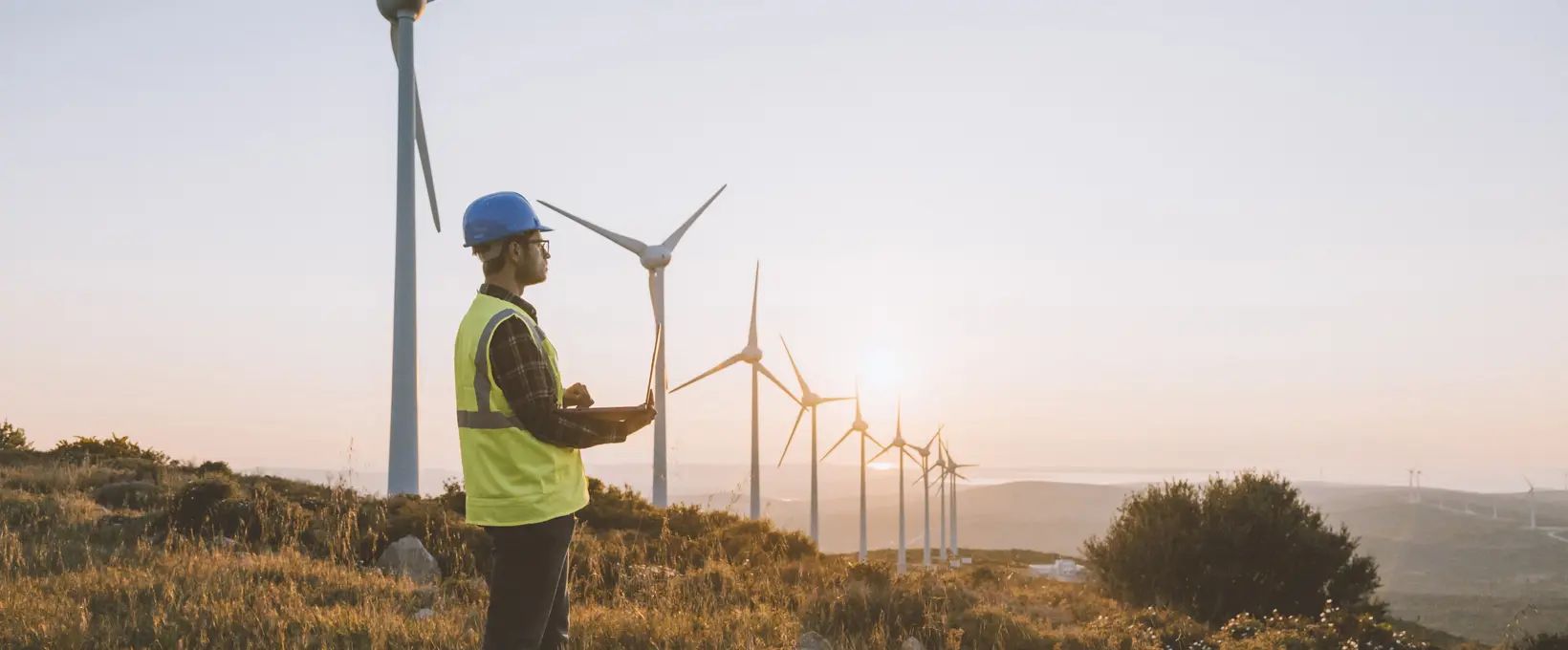 Man stood near windmills, on a hilltop wearing PPE and holding a clipboard